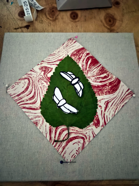 A linen looking fabric covered board with the white and pink square diamond paper from Lula in the center.  Pins at each corner are carefully keeping it in place without damaging the paper.  A large leaf sits in the center, stem down, with two paper cicada templates facing each other diagonally across it. The shadow box's frame is nowhere to be seen.