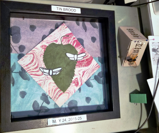 A black framed shadow box.  The square diamond of paper with white and pink swirls of color from Lula is in the center, and in the center of that is a large dried mulberry leaf, pointed downwards.  Two paper cicada templates are arranged on either side of the leaf, overlapping it.  They are offset from each other vertically as well, with the one on the right higher.  Off to the side, on the desk, you can see one of the cicadas pinned onto a scrap of cardboard.